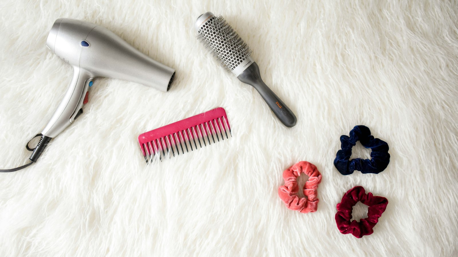A silver hair dryer, a round brush, a comb and scrunchies on a white rug