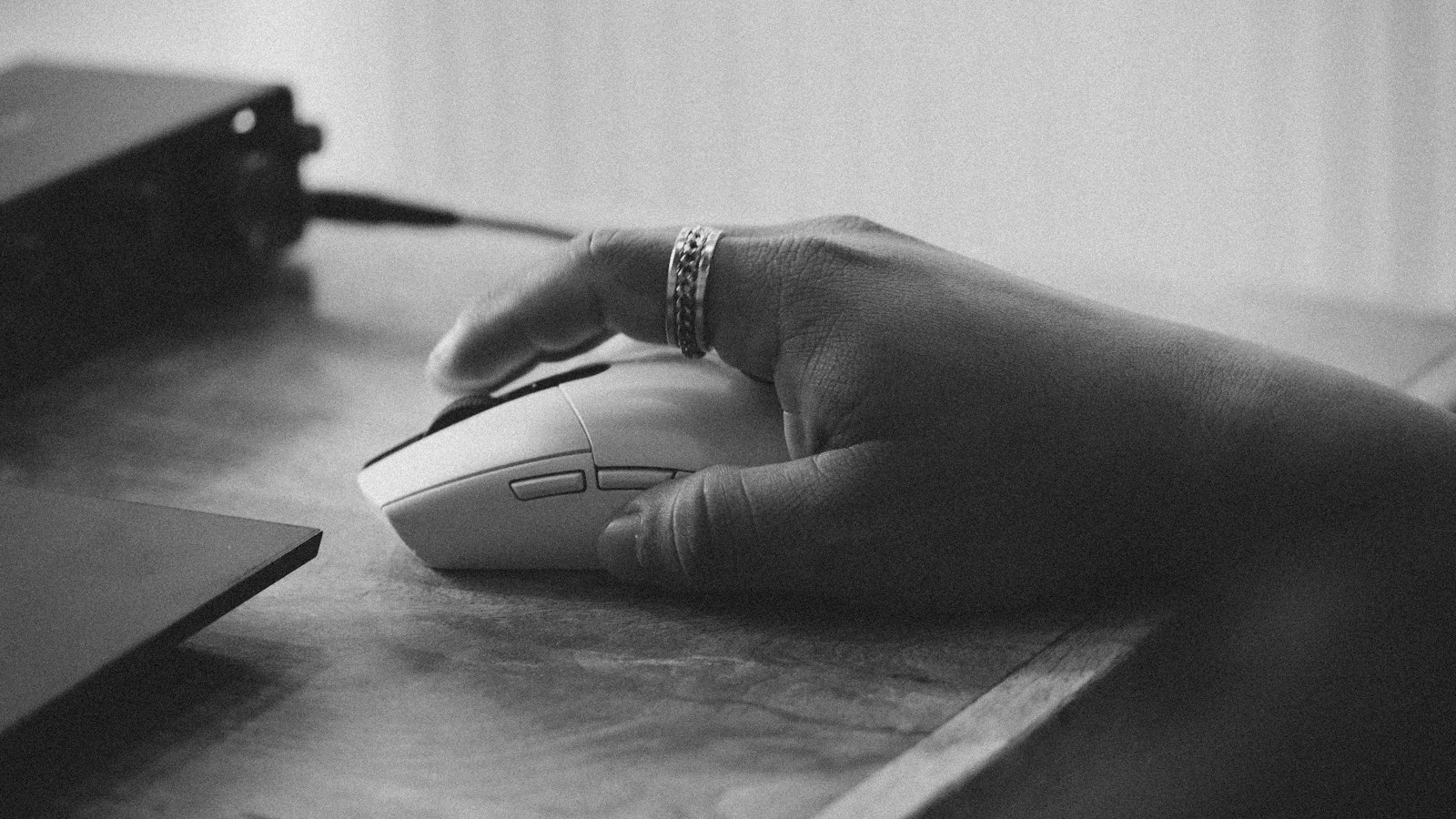 A hand resting on a computer mouse on a wooden desk, in black and white